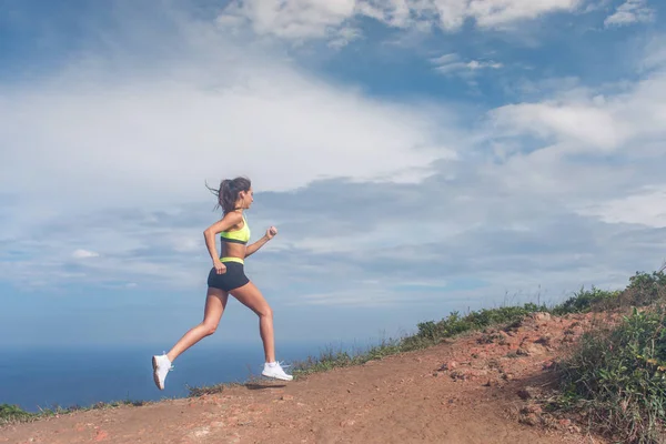 Mujer atlética corriendo por la montaña con el cielo y el mar de fondo. Corredor profesional haciendo ejercicio cardiovascular al aire libre en el paisaje natural —  Fotos de Stock