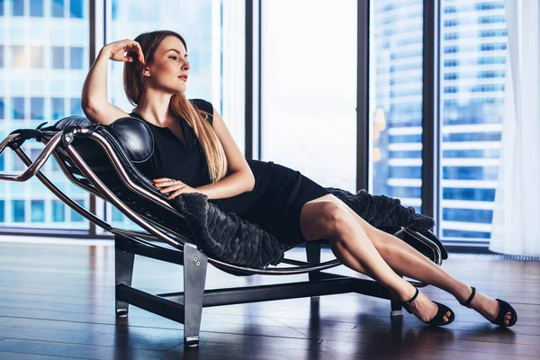 Modelo de moda con piernas largas y delgadas con vestido de cóctel negro tumbado en la silla del salón en el apartamento ático —  Fotos de Stock