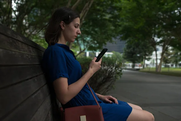 Vista lateral de una joven sentada en un banco en el parque sosteniendo su teléfono inteligente —  Fotos de Stock