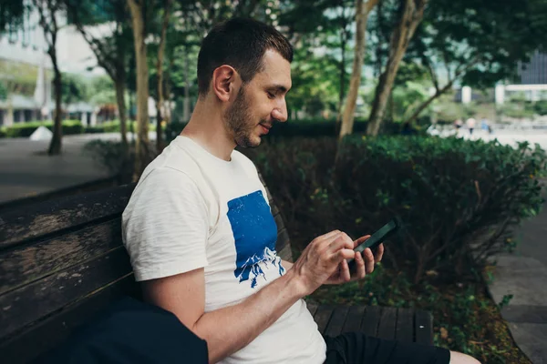 Muž sedí na lavičce v parku venku a používá smartphone — Stock fotografie