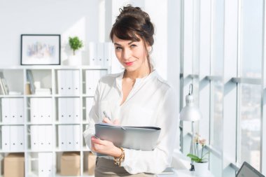 Kameraya, seyir modern ofiste ayakta, kağıt klasör, holding çekici iddialı işkadını gülümseyen.