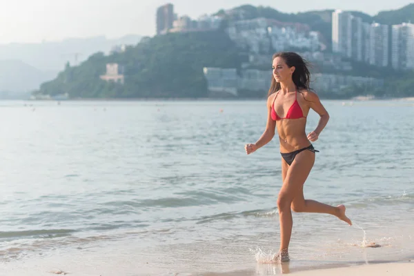 Sportieve jonge vrouw in bikini lopen langs het strand met fel zonlicht en bergachtige resort stad op achtergrond — Stockfoto