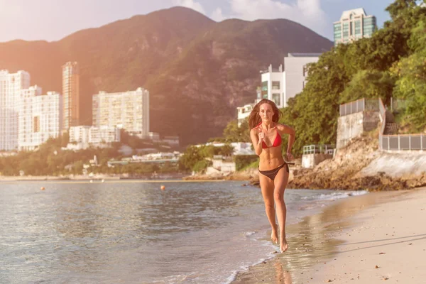 Adatta atleta donna che indossa bikini in esecuzione sulla spiaggia con sole splendente in camera e hotel resort colline sullo sfondo — Foto Stock