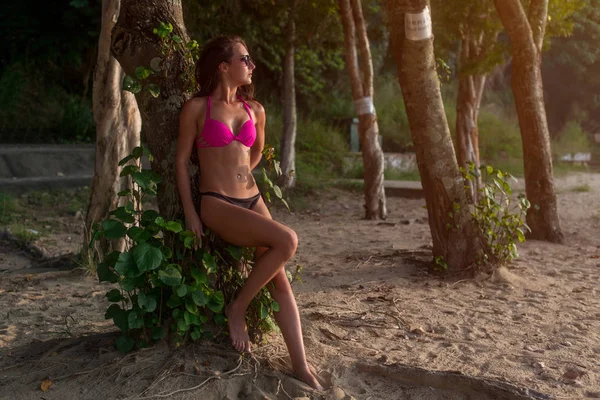 Fitness modèle brune femme portant un bikini et des lunettes de soleil appuyé contre l'arbre tenant ses cheveux sur la plage — Photo