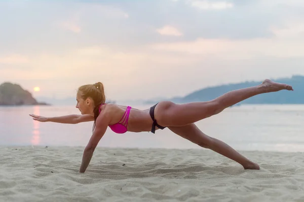 Jonge vrouw bij de uitoefening van de zwembroek op het strand dat zich uitstrekt van haar benen tijdens zonsondergang op zee. Fitness meisje doen oefeningen aan kust — Stockfoto