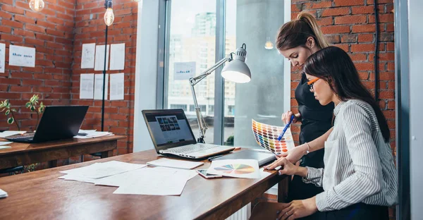 Mujeres jóvenes que trabajan en un nuevo diseño web utilizando muestras de color y bocetos sentados en el escritorio en la oficina moderna — Foto de Stock