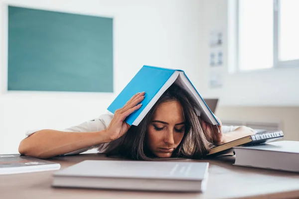 Estudiante cansada sentada a la mesa con un libro en la cabeza . — Foto de Stock