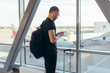 Havaalanında pencerenin yanında duran genç kadın elinde cep telefonuyla