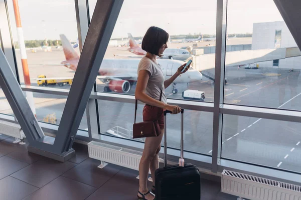 विमानतळ विंडो जवळ उभे तरुण स्त्री मोबाइल फोन धारण — स्टॉक फोटो, इमेज