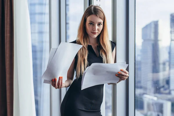 Woman holding documents, looking through papers, studying the report standing near window with view on skyscrapers — Stock Photo, Image