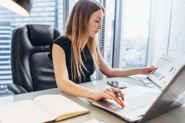 Diretor feminino trabalhando no escritório sentado na mesa analisando estatísticas de negócios segurando diagramas e gráficos usando laptop — Fotografia de Stock
