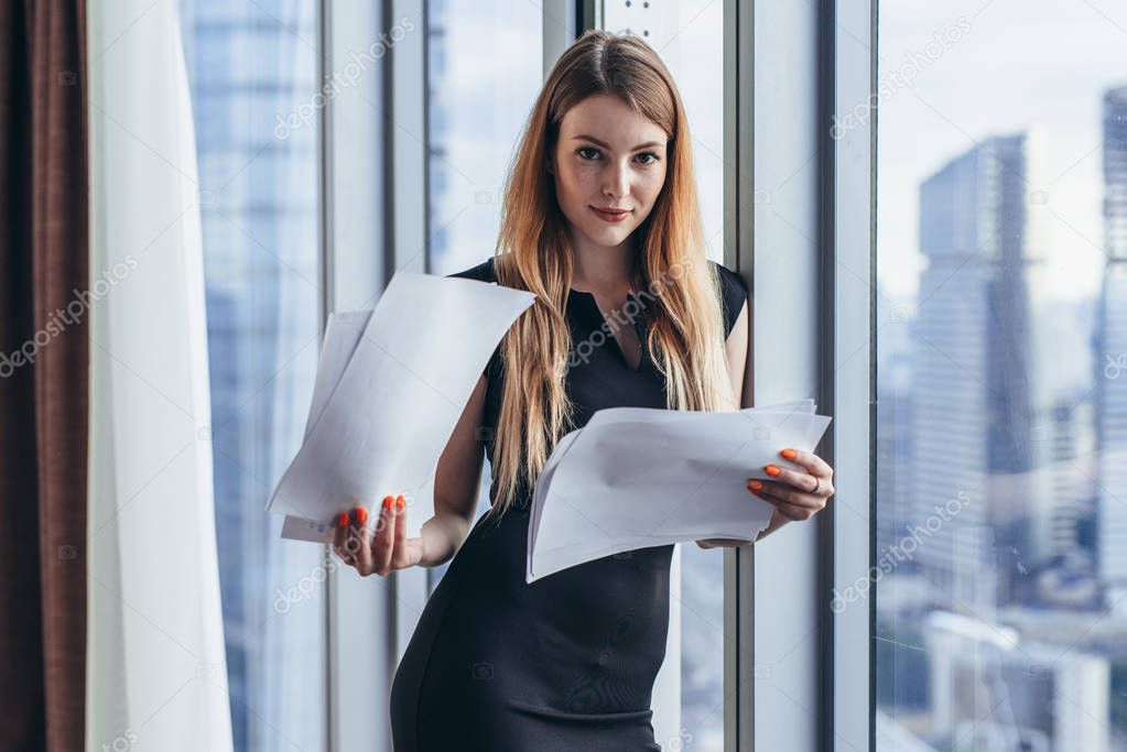 Woman holding documents, looking through papers, studying the report standing near window with view on skyscrapers