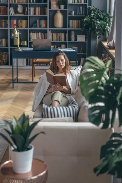 Jovem em casa está deitada no sofá com livro . — Fotografia de Stock