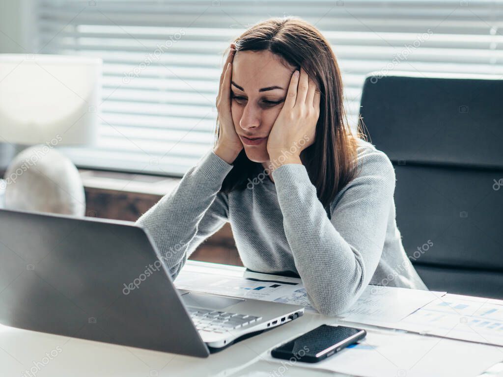 Woman sitting at table with her head leaning on her hands and looking at laptop monitor.