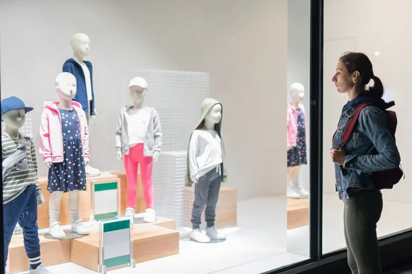 Kadın çocuk mağazasının önünde duruyor ve mankenlerin üzerindeki kıyafetlere bakıyor..