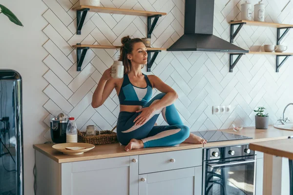 Femme sportive prenant le petit déjeuner dans la cuisine, fille assise sur la table et buvant — Photo