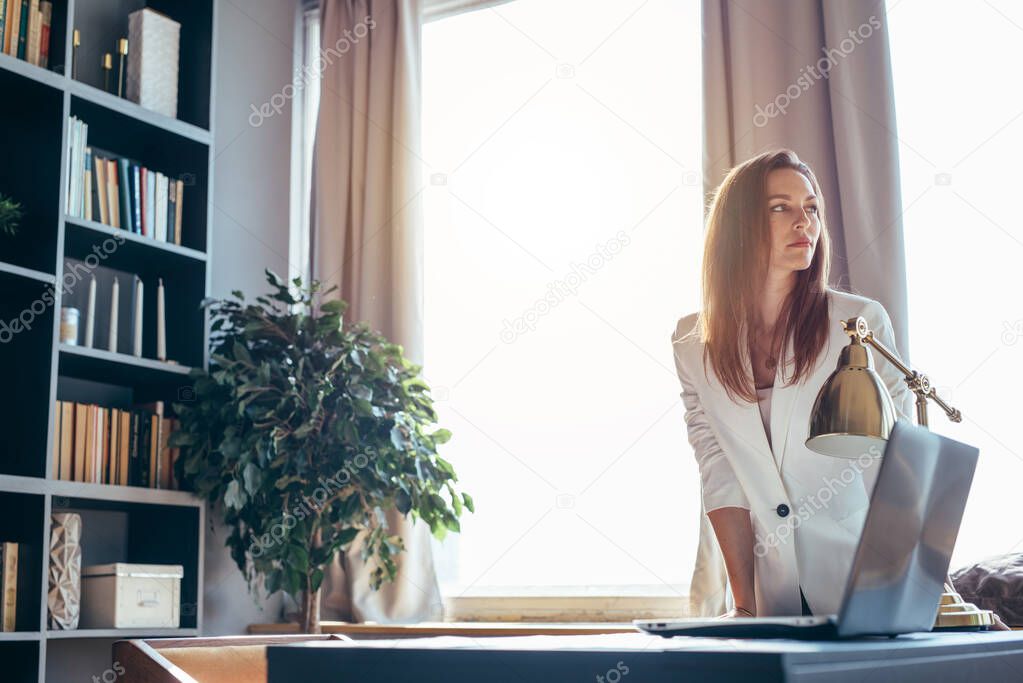 Business woman stands near the workplace leaning on the table.