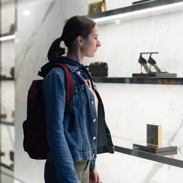 Egy fiatal nő áll a butik kirakatánál, táskákat és cipőket néz. — Stock Fotó