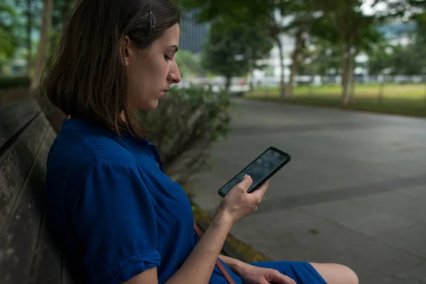 一个拿着智能手机坐在公园长椅上的女孩. — 图库照片