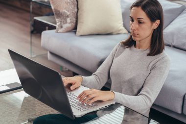 Genç bir kadın dizüstü bilgisayar kullanarak kanepede oturuyor..