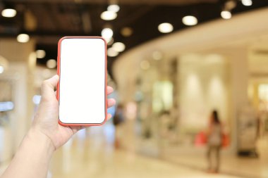 Alışveriş merkezinin arka planında akıllı telefon cihazı tutan ve ekranında beyaz fotokopi alanı olan bir adamın eli..