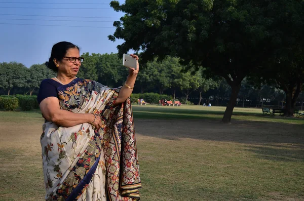 Old indian woman wearing a saree clicking a selfie in a park outdoor with a smartphone in Delhi, India