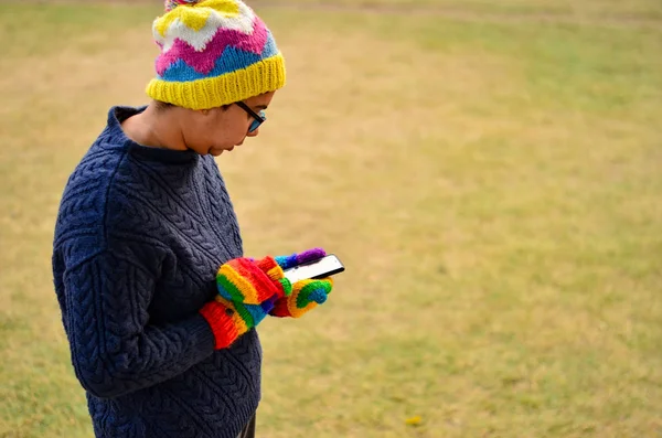 Woman wearing colorful mittens and cap operating phone in winters in a park in Delhi, India