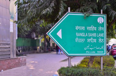 Central Delhi has unique street signs for road names. This photo shows the ubiquitous green direction board for Bangla Sahib Lane street sign Delhi, India written in English, Hindi, Punjabi and Urdu clipart