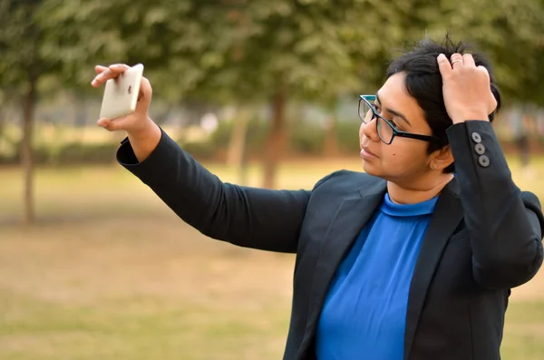 Young confident Indian professional woman checking her hair on her smartphone in an outdoor setting wearing a black business / formal suit with a blue top