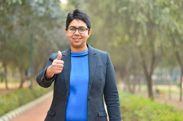 Portrait of a confident young Indian Corporate professional woman with short hair showing thumbs up sign (Camera focus on thumbs up) in an outdoor setting wearing a black business / formal suit