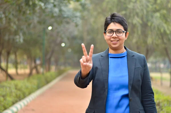Portrait of a confident young Indian Corporate professional woman with short hair showing Victory V sign (Camera focus on V) in an outdoor setting wearing a black business / formal suit