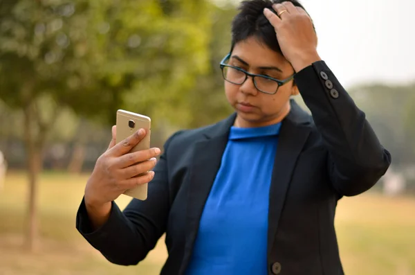 Young confident Indian professional woman checking her hair on her smartphone in an outdoor setting wearing a black business / formal suit with a blue top