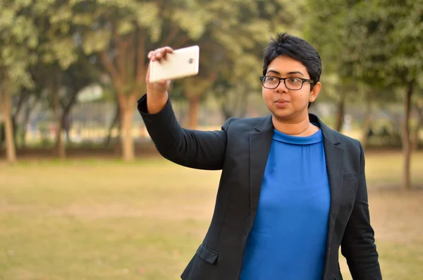 Young confident Indian Corporate professional woman  with short hair taking a selfie on her smartphone in an outdoor setting wearing a black business / formal suit with a blue top