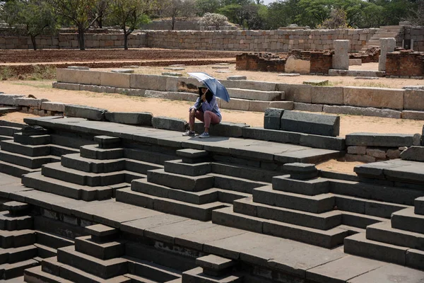 Female Indian tourist sitting with an umbrella at the public bath and the royal enclosure step water well in Hampi, Karnataka, India. Group of Monuments at Hampi is a UNESCO world heritage site.