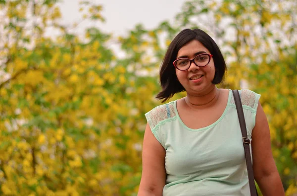 Young confident Indian woman wearing specs standing against yellow leaves during autumn weather with a sling bag on her shoulder in Delhi, India