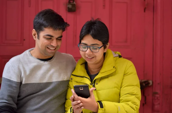 Young Indian working professional couple smiling against a pink door background. Lifestyle portrait shot of a young Indian family couple looking at their mobile phone and smiling