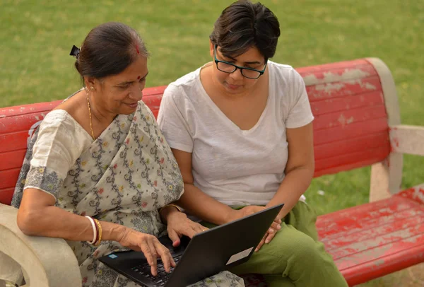 Young Indian girl helping an old Indian woman on a laptop sitting on a red bench in a park in Delhi, India