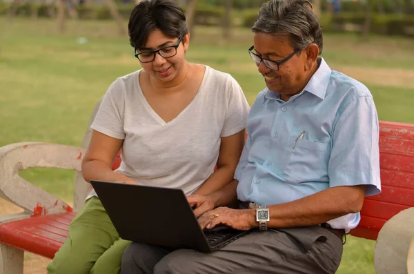 Young Indian woman helping an old Indian man on a laptop sitting on a red bench in a park in Delhi, India