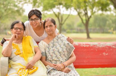 Delhi, Hindistan'da bir parkta kırmızı bir bankta oturan iki üst düzey Hintli anne ile Mutlu görünümlü genç Hintli kadın