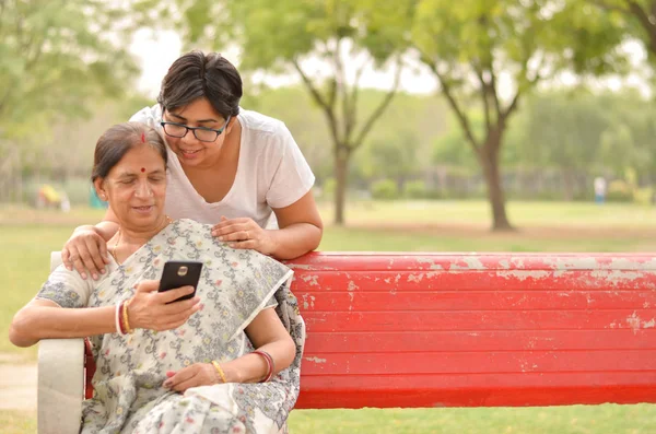 Young Indian girl with an old Indian woman looking at the mobile phone and busy talking sitting on a red bench in a park in Delhi, India