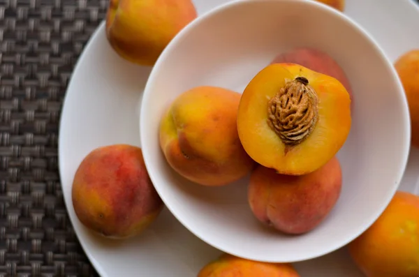 Peaches cut in half and full in a bone china white bowl kept on a white plate decorated on a brown table mat