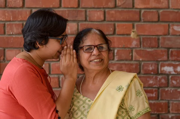 A senior and a young Indian woman mother daughter pair speaking softly into the other\'s ear, gossiping against a red brick wall in Delhi, India