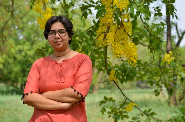 短い髪と眼鏡を持つ自信に満ちた若いインドの女性の肖像画は 黄色の花を持つ公園で伝統的な北インドのスーツドレスを着て屋外の設定で折り畳まれた手を渡った — ストック写真