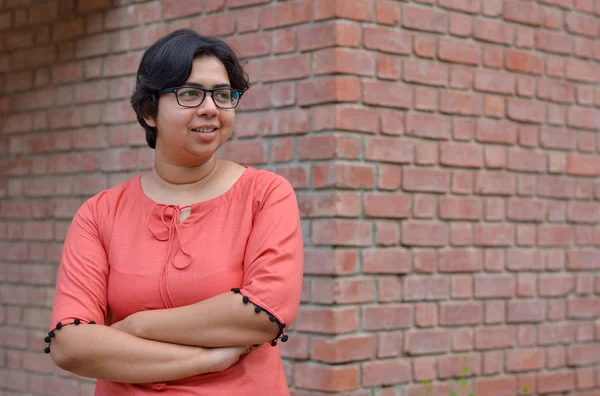 Portrait of a confident looking young Indian woman with short hair and spectacles, crossed folded hands in an outdoor setting wearing a traditional north Indian suit dress against a red brick wall