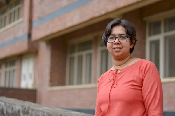 Portrait of a confident looking young Indian woman with short hair and spectacles, in an outdoor setting wearing a traditional north Indian suit dress against a red brick wall