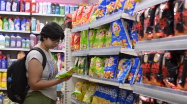 Delhi, Hindistan, 2019. Bir alışveriş merkezinde modern bir süpermarket mağazada sırasıra sergilenen cips / aperatifler bölümünde tarama genç bir kadının Video.