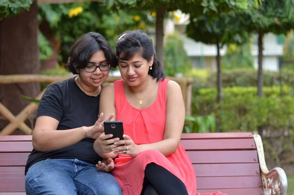 Twee Vrienden Zittend Een Rode Parkbank Kijkend Mobiele Telefoon Lachend — Stockfoto