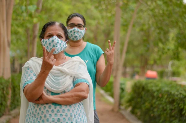 Close up Portrait of senior Indian lady and young lady wearing matching surgical cotton mask to protect themself from Corona Virus (COVID-19) pandemic showing V victory signs in a park in Delhi, India