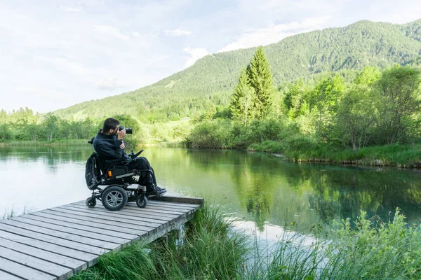 Hombre en silla de ruedas utilizando la cámara sin espejo cerca del lago en la naturaleza — Foto de Stock