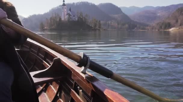 Movimento lento de remo remo em um barco no lago Bled com ilha Bled e igreja atrás dele e um reflexo no lago, com sol no barco — Vídeo de Stock